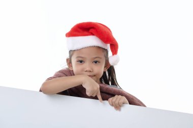 Noel Baba şapkası takan mutlu küçük bir kızın portresi beyaz arka planda izole edilmiş beyaz bir paneli işaret ediyor. Elinde boş bir Noel panosu var.