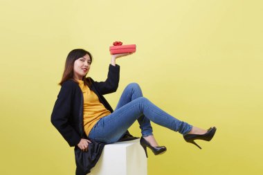 Tam boy portre. Elinde hediye tutan çekici genç kadın. Beyaz küp üzerinde izole edilmiş sarı arka plan.