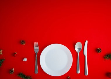 En düşük manzaralı Noel topları süslemesi ve kırmızı zemin üzerinde çatal, bıçak ve kaşık seti olan beyaz boş bir tabak.