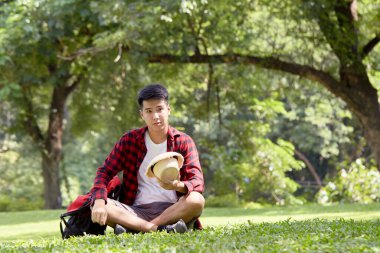 Genç Asyalı sırt çantalı adam yaz günü çimenlerde oturuyor yakışıklı adam tatilde açık hava yaşam tarzıyla eğleniyor.