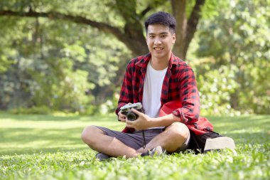 Genç Asyalı sırt çantalı adam yaz günü çimenlerde oturuyor yakışıklı adam tatilde açık hava yaşam tarzıyla eğleniyor.