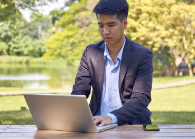 Takım elbiseli yakışıklı bir Asyalı iş adamı bahçede bir masada oturmuş akıllı bir telefon ve dizüstü bilgisayarla çalışıyor..