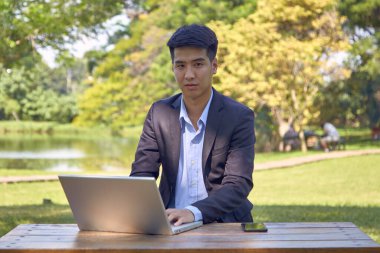 Takım elbiseli yakışıklı bir Asyalı iş adamı bahçede bir masada oturmuş akıllı bir telefon ve dizüstü bilgisayarla çalışıyor..
