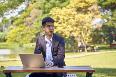 Takım elbiseli yakışıklı bir Asyalı iş adamı bahçede bir masada oturmuş akıllı bir telefon ve dizüstü bilgisayarla çalışıyor..