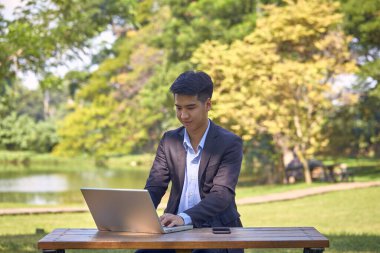 Takım elbiseli yakışıklı bir Asyalı iş adamı bahçede bir masada oturmuş akıllı bir telefon ve dizüstü bilgisayarla çalışıyor..