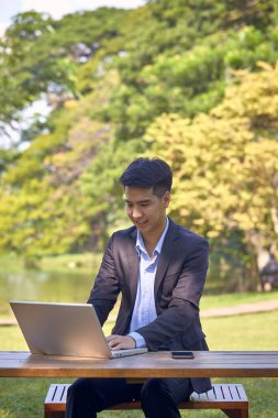 Takım elbiseli yakışıklı bir Asyalı iş adamı bahçede bir masada oturmuş akıllı bir telefon ve dizüstü bilgisayarla çalışıyor..