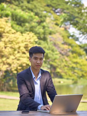 Takım elbiseli yakışıklı bir Asyalı iş adamı bahçede bir masada oturmuş akıllı bir telefon ve dizüstü bilgisayarla çalışıyor..