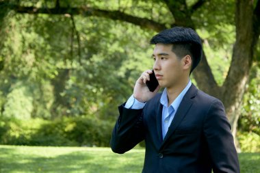 Genç Asyalı iş adamı akıllı bir telefonla parkta yürüyor..
