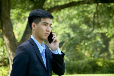 Genç Asyalı iş adamı akıllı bir telefonla parkta yürüyor..