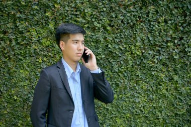 Genç Asyalı iş adamı akıllı bir telefonla parkta yürüyor..