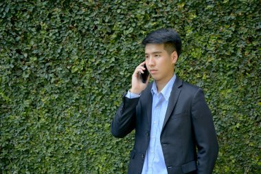 Genç Asyalı iş adamı akıllı bir telefonla parkta yürüyor..