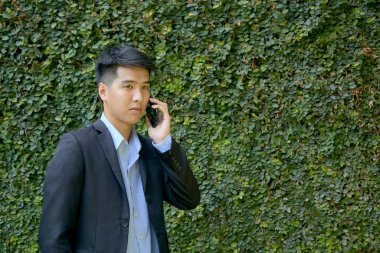 Genç Asyalı iş adamı akıllı bir telefonla parkta yürüyor..