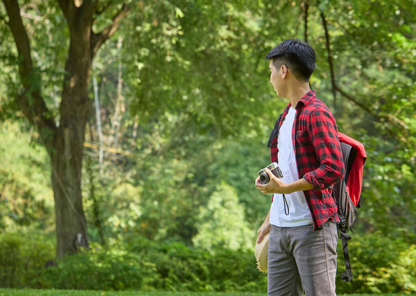 A young Asian backpacker holding a camera standing in a nature park.