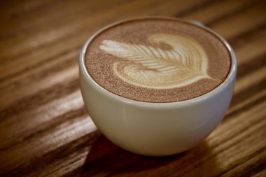 Yüzeyinde süt sanatı olan bir fincan sıcak kahve, ahşap masaya konur.