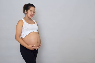 Gülümseyen hamile Asyalı kadın gri arka planda karnına dokunuyor.