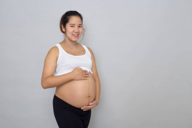 Gülümseyen hamile Asyalı kadın gri arka planda karnına dokunuyor.