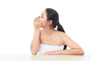 Güzel bir Asyalı kadın kameraya bakıyor ve küçük bir kozmetik fırçasıyla beyaz arka planda çıplak omuzlarıyla kaşının resmini çizmek için elini kaldırdı.