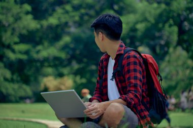 Yakışıklı Asyalı bir adam bahçede dizüstü bilgisayar kullanarak oturuyor.