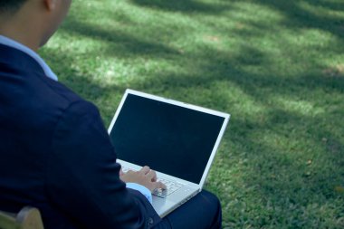 Parkta dizüstü bilgisayarı ve dizüstü bilgisayarı olan Asyalı genç bir işadamı bahçede otururken dizüstü bilgisayarla görüntülü konuşma yapıyor. İş ve teknoloji