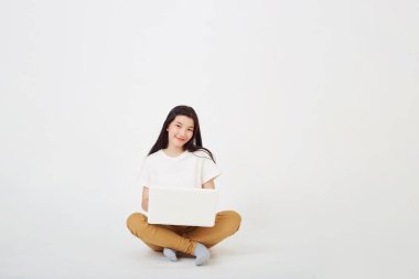 Okul konseptine geri dönersek, Mutlu Asyalı genç bir kadın yerde bacak bacak bacak üstüne atmış bir şekilde oturuyor ve gri arka planda dizüstü bilgisayar kullanıyor.