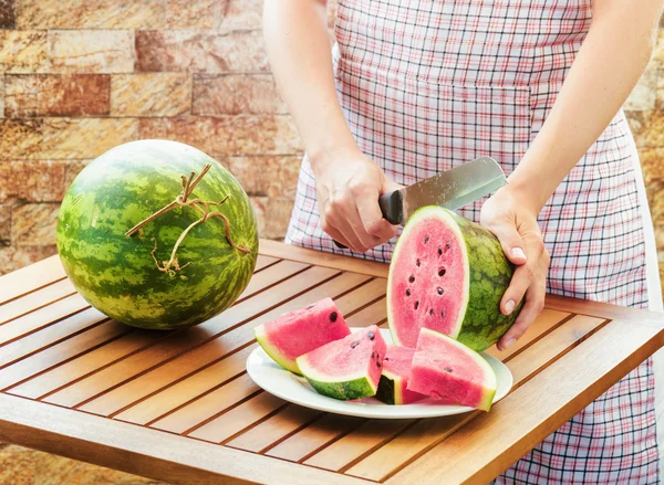 Young woman in apron slicing watermelon on wooden table — Stock Photo, Image