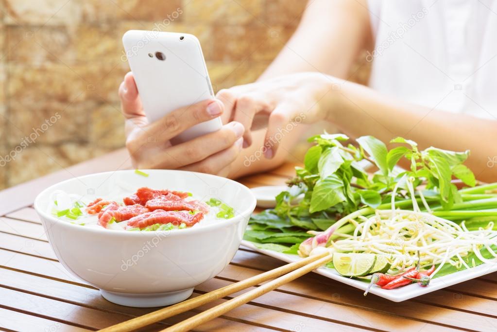 Young woman eating the Pho Bo and using a smartphone in Vietnam