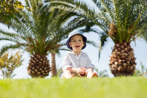 Glücklicher kleiner Junge liegt auf grünem Gras unter Palmen — Stockfoto