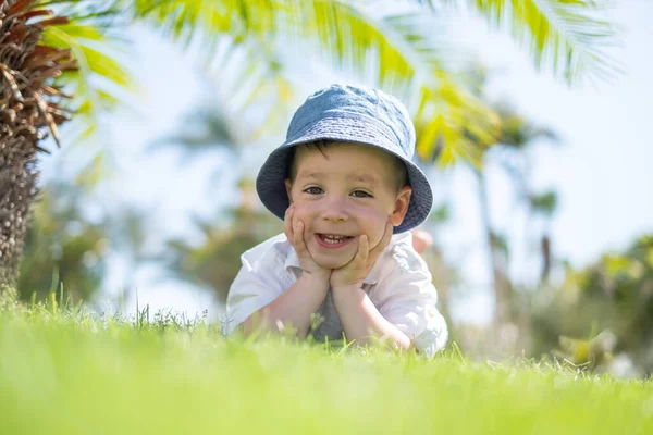 Glücklicher kleiner Junge liegt auf grünem Gras unter Palmen — Stockfoto