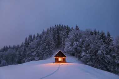 Kış manzarası. Gizemli bir gece. Bahçedeki eski ahşap kulübe karla kaplıydı. Lambalar akşam vakti evi aydınlatıyor. Dağlar ve ormanlar. Duvar kağıdı arkaplanı.