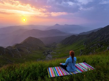 Bahar sabahında gün doğumu. Yogacı kız yüksek dağlardaki çimlerde meditasyonda çizgili minderde oturuyor ve gökyüzü ışınlar ve bulutlarla kaplı. Duvar kağıdı arka planı. Doğal manzara..