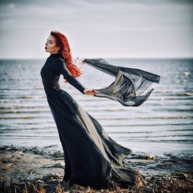 Beautiful sad goth girl with cloth in hands standing on sea shore
