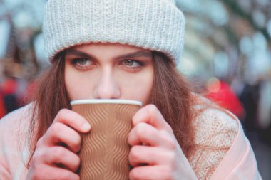 Winter holidays, christmas, hot drinks and people concept. Happy beautiful young woman in warm clothes drinking coffee from disposable paper cups in old town