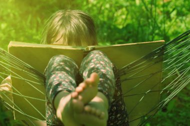 Genç kız rahat bir hamakta yeşil çimlere karşı kitap okuyor.