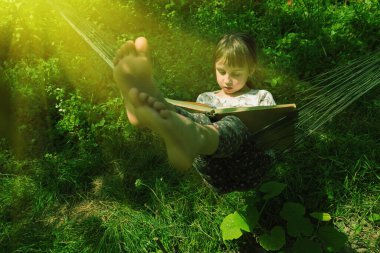 Genç, güzel bir kız, hamakta kitap okumaktan zevk alıyor. Dışarıda, güneşin altında yeşil bahçede. Yatay resim.