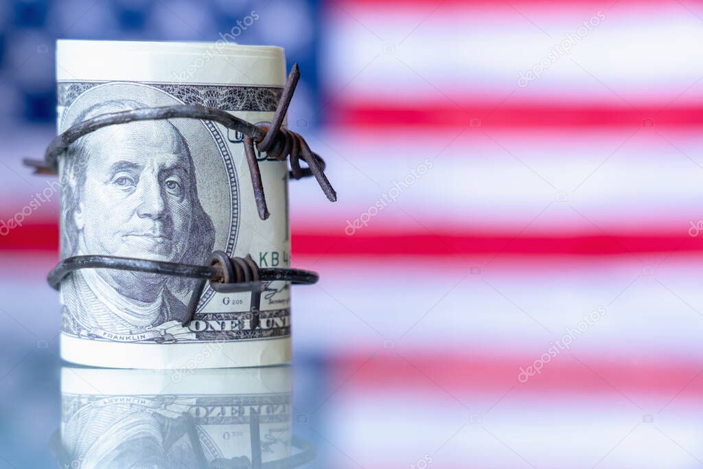 US Dollar currency wrapped in barbed wire against flag of United States as symbol of economic warfare, sanctions and embargo busting concept. Copy space for design or text.
