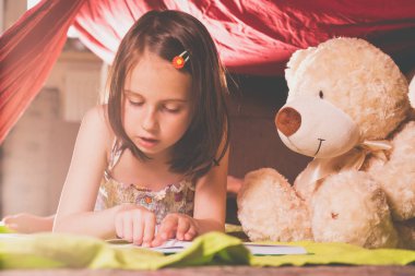 Genç güzel kız büyük bir kitap okuyor. Yere yakın bir yerde bir oyuncak ayı oturuyor. Rahatlama ve dostluk kavramı