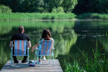 Göldeki ahşap rıhtımdaki çift. Gölün etrafında yeşil bir orman ve dağlar var. Dinlenme, seyahat, yaz ve tatil konsepti. Yatay resim.