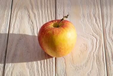 Bir kırmızı ve sarı elma, tahtalardan yapılmış açık ahşap bir masa üzerinde, güneş tarafından gölgelerle aydınlatılmış, doğal desenli.