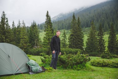 Arka planda dağ manzaralı çadırın yanında kalan genç bir adam.