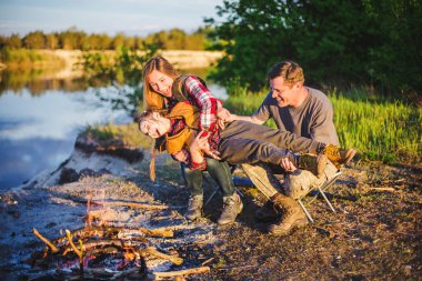 Genç bir aile gün batımında kamp ateşinin önünde bir çocukla geziyor ve kamp yapıyor. Arka planda bir nehir var.