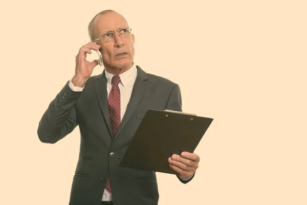Studioaufnahme eines leitenden Geschäftsmannes, der Klemmbrett hält, während er mit dem Handy spricht und denkt — Stockfoto