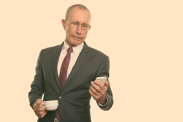 Studio skott av senior affärsman som håller kaffekopp medan du använder mobiltelefon — Stockfoto