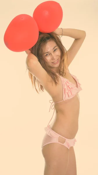 Studio împușcat de tânără femeie asiatică fericită zâmbind în timp ce ține două baloane roșii pe cap și poartă lenjerie intimă — Fotografie, imagine de stoc