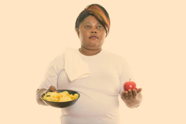Stúdió lövés kövér fekete afrikai nő gondolkodás közben gazdaság tál burgonya chips és piros alma — Stock Fotó