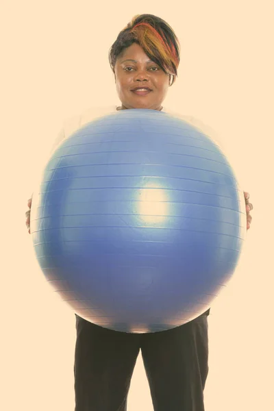 Studio skott av glad fett svart afrikansk kvinna ler och står medan du håller träningsboll redo för gym — Stockfoto