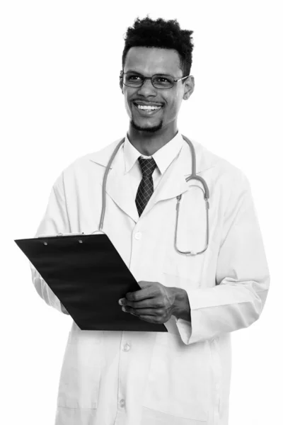 Young happy African man doctor smiling while holding clipboard and thinking — Stock Photo, Image