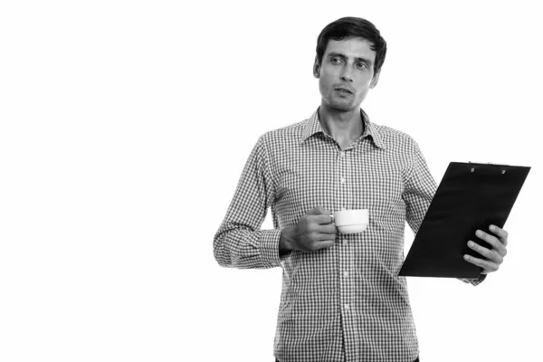 Studio shot of young handsome businessman thinking while holding coffee cup and clipboard — Stock Photo, Image