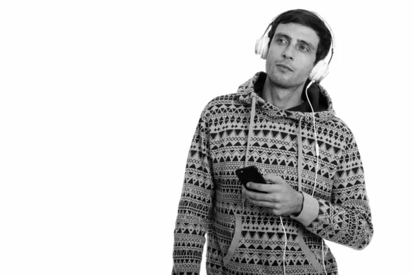 Studio shot of young handsome man listening to music while thinking and holding mobile phone — Stock Photo, Image
