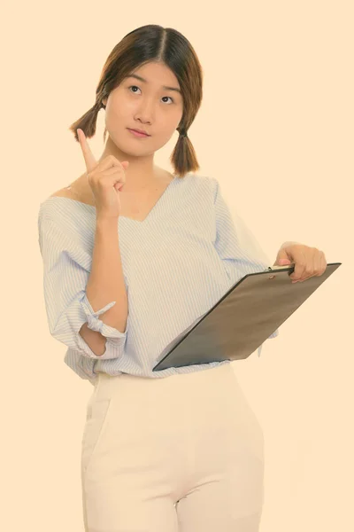 Joven mujer asiática reflexiva apuntando con el dedo hacia arriba mientras sostiene el portapapeles —  Fotos de Stock