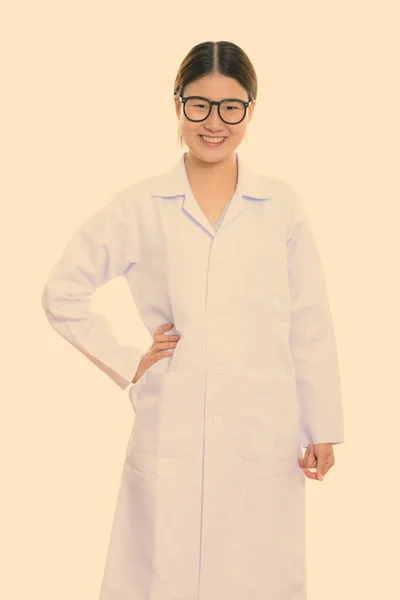 Estúdio tiro de jovem feliz asiático mulher médico sorrindo com a mão no quadril — Fotografia de Stock